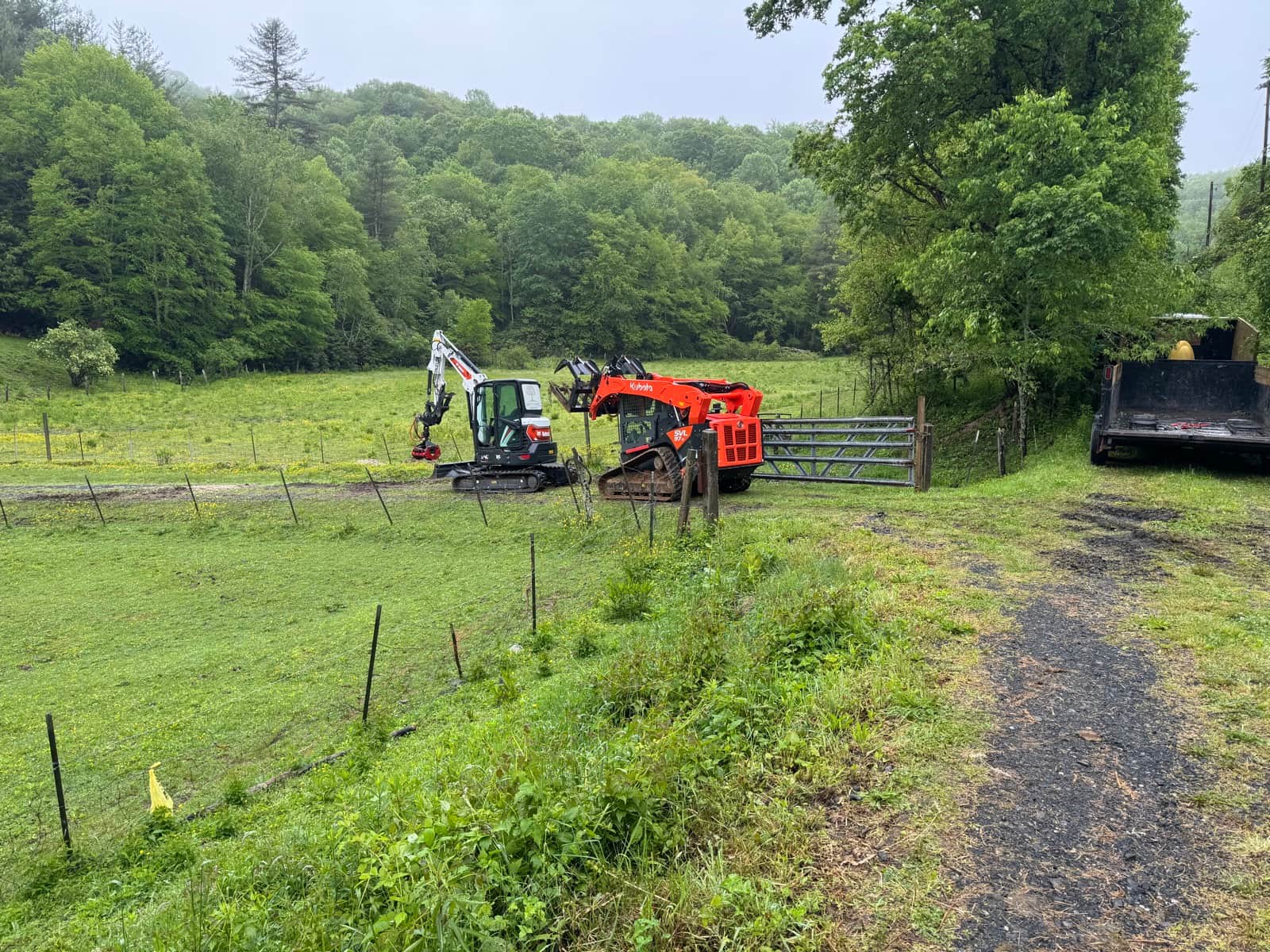 Excavator and Kubota working in a green field with wooded mountains during land clearing